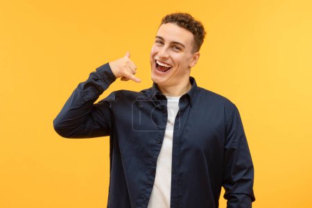 Photo for Hey you. Happy playful handsome young guy in casual showing call me gesture and smiling, isolated on yellow studio background, flirting, enjoying good day - Royalty Free Image