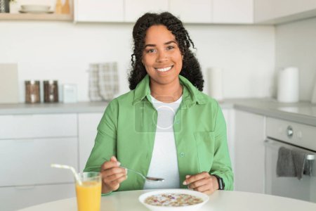 Foto de Feliz dama negra saboreando el desayuno, sentada con un plato de cereales ante ella y un vaso de jugo de naranja, disfrutando de una comida y una dieta equilibrada en la acogedora cocina de su casa. Nutrición y bienestar - Imagen libre de derechos