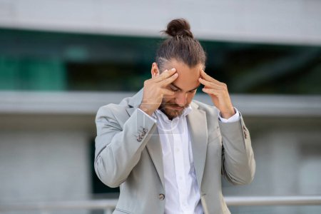 Photo for Closeup of stressful young businessman standing by office building, touching his head, upset male entrepreneur suffering from problems at job, having business failure, thinking about solution - Royalty Free Image