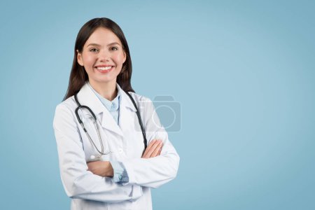 Selbstbewusste junge kaukasische Ärztin in weißem Mantel mit Stethoskop, die Arme vor blauem Hintergrund verschränkt, freundlich guckend, freier Raum
