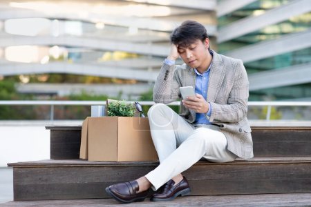 Téléchargez les photos : Asiatique homme d'affaires spécialiste en utilisant smartphone assis au chômage après le licenciement sur la rue de la ville urbaine en plein air, face à la mise à pied. Crise économique, victime du concept de réduction des effectifs des entreprises - en image libre de droit