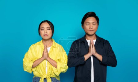 Photo for Serious pensive asian man and lady in casual with closed eyes pray, dream, make wish come true, isolated on blue studio background. Expects good news and luck, hope and belief - Royalty Free Image