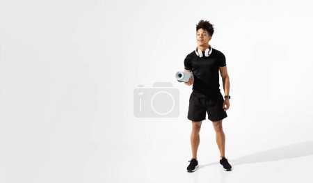 Photo for Athletic young latin man smiles with wireless headphones on neck, holding fitness mat while standing ready for gym workout on white studio backdrop. Free space for sport advertisement - Royalty Free Image
