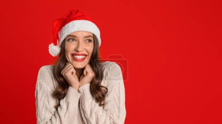 Téléchargez les photos : Dame européenne souriante au chapeau de Père Noël regarde de côté anticipant les festivités, sur fond de studio rouge, incarnant la joie de Noël et la saison du Nouvel An. Panorama, espace de copie - en image libre de droit