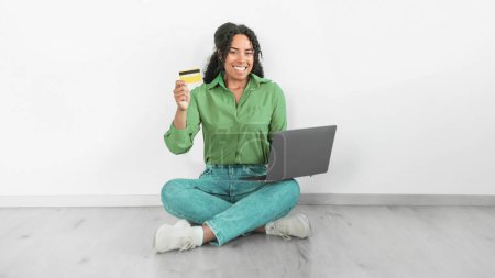 Photo for Smiling Black woman using laptop and credit card for financial transaction, symbolizing the ease of online payments and shopping, sitting on floor over white wall indoor. Ecommerce offers. Panorama - Royalty Free Image