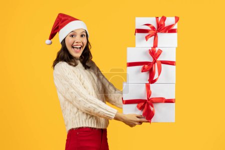 Téléchargez les photos : Femme caucasienne souriante dans le chapeau de Père Noël posant avec pile de cadeaux de Noël, tenant des boîtes blanches enveloppées avec des noeuds de ruban rouge, debout sur fond de studio jaune. Bannière de vente de vacances de Noël - en image libre de droit