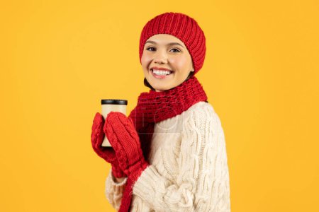 Téléchargez les photos : Happy lady tient une tasse de café chaud en souriant tandis qu'elle boit debout sur fond de studio jaune, posant avec une tasse en papier portant des tricots rouges d'hiver. Nouvel An concept d'ambiance de vacances - en image libre de droit
