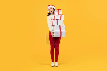 Photo for Joyful woman poses with stack of many Xmas festive boxes on yellow studio background, offering Christmas gifts greeting and smiling to camera. Full length. New Year sale advertisement - Royalty Free Image