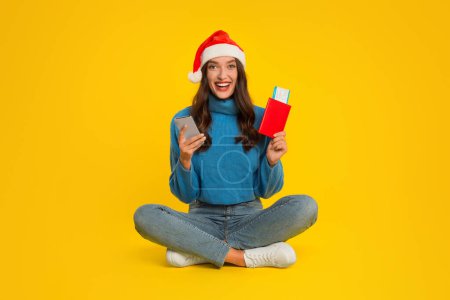 Téléchargez les photos : Vacances de Noël. Joyeux jeune femme globetrotter dans le chapeau Santa détient téléphone et billets d'avion dans le passeport, réserve son escapade de Noël, assis sur fond de studio jaune - en image libre de droit