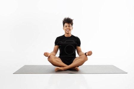 Foto de Joven chico de fitness en fitwear practica yoga sentado en posición de loto en la estera del gimnasio, meditando y relajándose en el fondo blanco del estudio. Hombre deportivo sonríe y disfruta de la calma durante la meditación de entrenamiento - Imagen libre de derechos