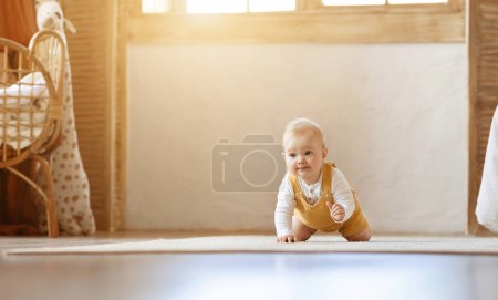 Téléchargez les photos : Adorable bébé garçon blond heureux rampant sur le sol par la maison dans le salon. Portrait d'enfant tout-petit mignon souriant se déplaçant seul par maison, regardant l'espace de copie et souriant - en image libre de droit