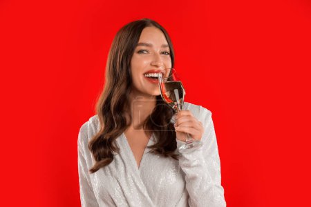 Téléchargez les photos : Joyeuse jeune femme élégante appréciant le goût de la boisson au champagne, posant avec un verre de vin mousseux et souriant à la caméra tout en célébrant les vacances, fond de studio rouge. Portrait tourné - en image libre de droit