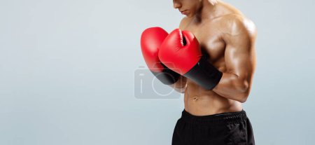 Photo for Unrecognizable muscular boxer man standing shirtless with red gloves on hands, posing in strong confident stance, on gray studio backdrop with free space, showcasing fighting spirit. Panorama - Royalty Free Image