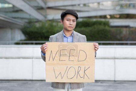 Téléchargez les photos : Homme de bureau coréen sans emploi montre Besoin affiche de papier de travail, regardant la caméra avec une expression malheureuse, debout en plein air du centre d'affaires. Chômage et réduction des effectifs des entreprises - en image libre de droit