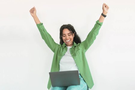 Photo for Joyful Black woman with laptop on lap cheers success, celebrating triumph in online freelance career, sitting against white studio background. Freelancer rejoices in front of her computer - Royalty Free Image