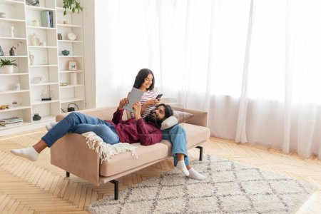 Foto de Gadget adicción, las tecnologías modernas y el estilo de vida de los millennials. Vista de ángulo alto de los cónyuges indios jóvenes pareja descansando en el sofá en casa, utilizando tableta digital y teléfono inteligente, espacio de copia - Imagen libre de derechos