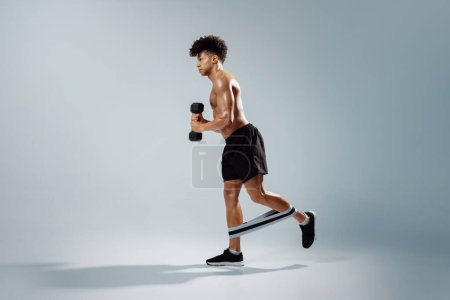 Foto de Determinado joven atleta sosteniendo pesas mientras hace ejercicio con banda de goma en las piernas, de pie sin camisa sobre fondo gris, destacando los brazos musculares y el torso. Gimnasio entrenamiento y fitness - Imagen libre de derechos