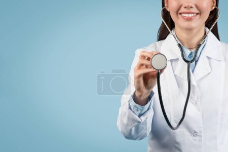 Téléchargez les photos : Jeune médecin européenne en uniforme médical blanc effectue un examen médical à l'aide d'un stéthoscope, sur fond bleu, gros plan, recadré, espace libre - en image libre de droit