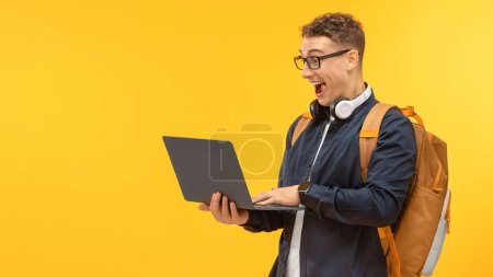 Téléchargez les photos : Positif jeune homme joyeux dans un étudiant occasionnel avec sac à dos et ordinateur portable PC dans ses mains posant isolé sur fond de studio jaune, bannière pour le concept d'éducation en ligne - en image libre de droit
