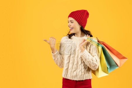 Photo for Cheerful woman with shopping bags in cozy knit sweater points thumb aside to empty space for winter seasonal offers, on yellow studio background. Christmas sales concept - Royalty Free Image
