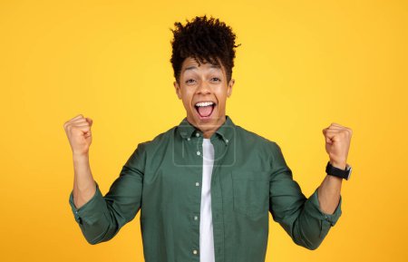 Téléchargez les photos : Jeune homme afro-américain excité avec une coiffure élégante serrant les poings geste, levant la main, célébrant le succès isolé sur fond de studio jaune - en image libre de droit