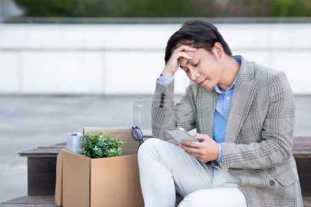 Photo for Fired japanese office worker guy holds his mobile phone sitting unhappily outdoor in urban area, experiencing uncertainty of unemployment, reflecting the tough job market - Royalty Free Image