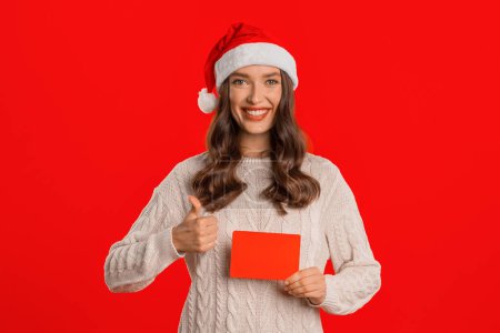 Téléchargez les photos : Femme européenne dans le chapeau de Père Noël donne pouces jusqu'à des ventes de vacances, tenant la carte-cadeau dans un décor de studio rouge festive. Acheteur dame annonce rabais d'hiver, présentation certificat d'achat - en image libre de droit