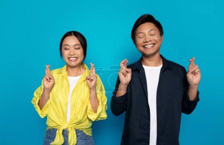 Photo for Happy pensive asian couple in casual with crossed fingers dream, make wish come true, isolated on blue studio background. Emotions think, sales, expects good news and luck - Royalty Free Image