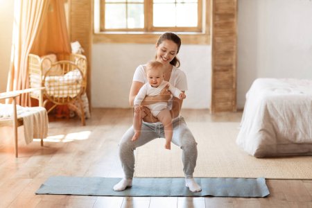 Photo for Sport activities with baby. Young mom exercising with her infant child at home. Happy sporty woman holding toddler son in her arms while doing squats training on fitness mat in bedroom, copy space - Royalty Free Image