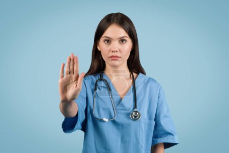 Téléchargez les photos : Jeune infirmière européenne dévouée dans des signaux uniformes médicaux bleus s'arrêtent avec confiance sur fond bleu, véhiculant un message de santé - en image libre de droit