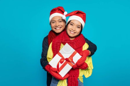 Téléchargez les photos : Heureux homme japonais millénaire dans les câlins chapeau femme avec boîte cadeau, profiter des vacances d'hiver, cadeaux de Noël, isolé sur fond de studio bleu. Week-end, froid et célébration du Nouvel An ensemble - en image libre de droit