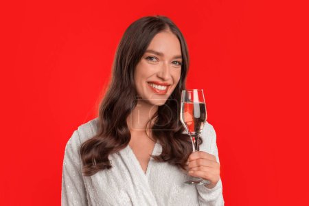 Foto de ¡Salud! Retrato de una joven mujer caucásica en vestido lujoso ofreciendo tostadas de champán en un estudio rojo, sosteniendo una copa de vino espumoso cerca de la cara, encarnando la celebración festiva en Navidad. - Imagen libre de derechos