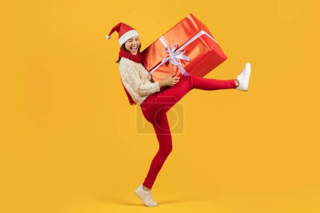 Foto de Entrega de regalos de Navidad. Jovencita divertida que lleva una gran caja de regalo envuelta en rojo caminando sobre el fondo del estudio amarillo, simbolizando la alegría y el espíritu de los regalos de Navidad que dan. Disparo completo - Imagen libre de derechos