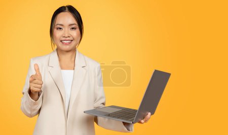 Photo for Glad confident young korean business lady in suit, with computer, make thumb up gesture, isolated on orange studio background. Work, study and business approve, recommendation sign - Royalty Free Image
