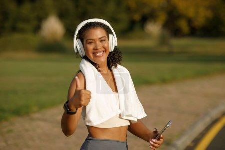 Téléchargez les photos : Femme de fitness brésilienne écouter de la musique dans les écouteurs par téléphone le matin courir à l'extérieur dans le parc public, geste pouces levés, approuver un mode de vie actif et sain - en image libre de droit