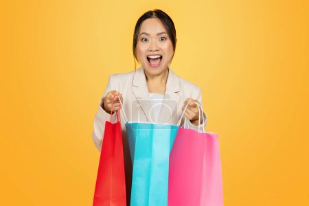 Téléchargez les photos : Étonné heureux jeune japonaise dame shopaholic ouvert sacs colorés, avec des achats profiter du shopping, isolé sur fond de studio orange. Grande vente, style de vie et nouvelle mode - en image libre de droit