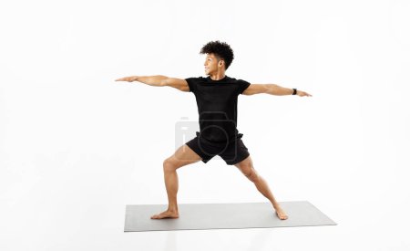 Photo for Brazilian fitness instructor guy demonstrating yoga warrior pose, balanced and strong on mat, exercising wearing fitwear, promoting wellness and healthy lifestyle, on white studio background - Royalty Free Image