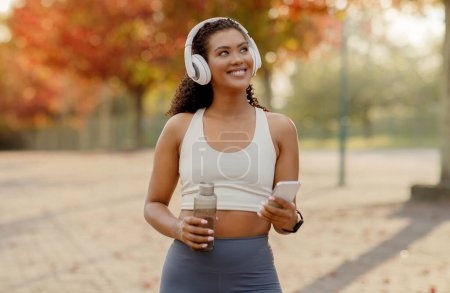 Téléchargez les photos : Jeune femme sportive écoutant de la musique au téléphone avec écouteurs, tenant une bouteille d'eau debout à l'extérieur, prête pour le jogging matinal ou l'entraînement de fitness dans un parc urbain - en image libre de droit