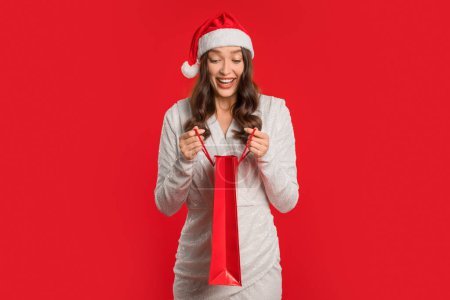 Téléchargez les photos : Shopping du Nouvel An. Millennial lady in Santa pose chapeau avec sac à provisions, jetant un coup d'oeil à l'intérieur de la recherche cadeau contre vibrant cadre studio rouge. Acheteur excité tenant des achats de Noël. - en image libre de droit