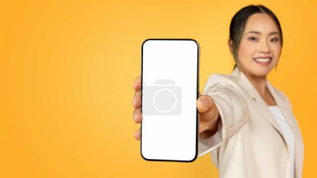 Photo for Cheerful young korean lady in suit, show mobile phone with empty screen, enjoy sale, ad and offer, isolated on orange studio background, panorama. Recommendation for work, study and business app - Royalty Free Image