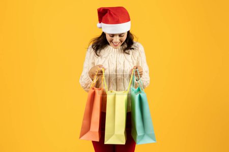 Photo for Young woman clutching colorful paper shopping bags wearing Santa hat, exudes joy against yellow studio backdrop, epitomizing festive spirit of Christmas commerce and sales season - Royalty Free Image