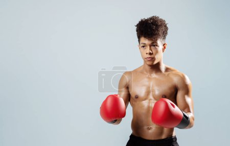 Photo for Muscular latin boxer guy posing shirtless with red gloves in fighting stance, expressing athletic strength and motivation, standing on gray studio backdrop with copy space - Royalty Free Image