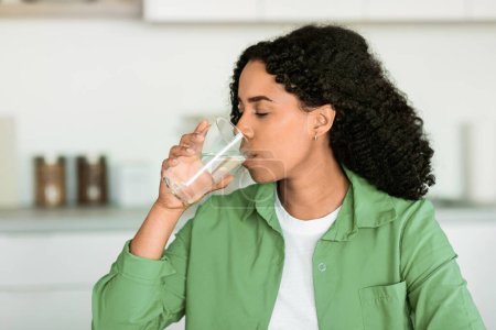 Photo for Black woman enjoys refreshing glass of water in her kitchen indoors, reflecting healthy lifestyle, hydration and balanced diet for weight loss health. African American lady sips mineral water - Royalty Free Image