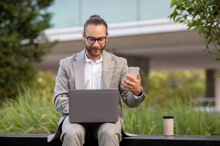 Photo for Modern Technologies For Business. Young Businessman With Laptop And Smartphone Sitting On Bench Outdoors, Handsome Male Entrepreneur In Suit Working Remotely Outside, Enjoying Multitasking - Royalty Free Image