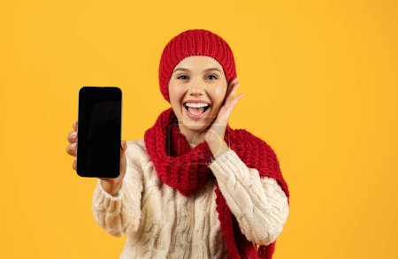 Foto de Joven alegre en ropa de punto de invierno muestra el teléfono con pantalla en blanco, maqueta para las promociones de la aplicación de Año Nuevo contra el fondo del estudio amarillo, la aplicación de publicidad y las compras navideñas modernas - Imagen libre de derechos