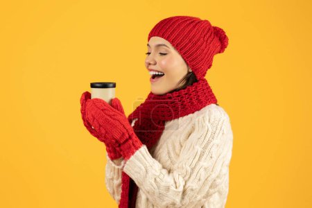 Foto de Mujer joven emocionado disfruta acogedor momento de invierno con café, sosteniendo taza de papel, vestido de punto rojo, sobre fondo de estudio amarillo. Vacaciones bebidas temporada y ofertas calientes concepto - Imagen libre de derechos