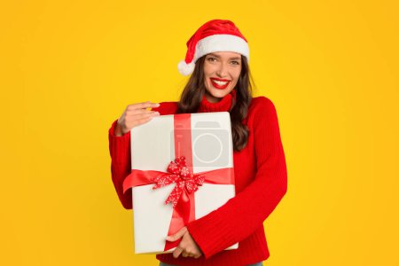 Foto de La mujer europea abraza alegremente la caja de regalo envuelta en un fondo de estudio amarillo, sonriendo a la cámara mientras posa con el regalo de Navidad con lazo de cinta roja. Ideal para promover las ventas y ofertas navideñas - Imagen libre de derechos