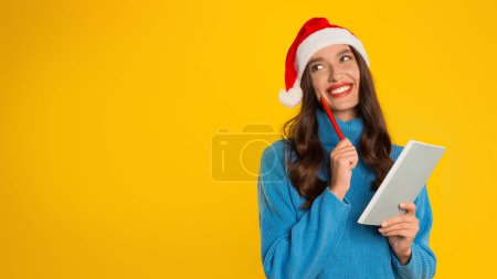 Téléchargez les photos : Jeune femme rêveuse en pull festif et chapeau de Père Noël écrit sa liste de souhaits de Noël dans son journal intime, tenant un cahier et un stylo sur fond jaune. Espérance et projets de vacances. Panorama, espace de copie - en image libre de droit