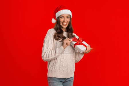 Photo for Christmas Surprise. Young Woman Holding Gift Box And Smiling To Camera, Posing for Festive Advertisement Against Red Studio Backdrop. Merry Xmas, Presents Delivery - Royalty Free Image