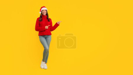 Téléchargez les photos : Joyeux jeune femme dans le chapeau Santa pointe de côté pour inviter l'espace libre, parfait pour les promotions festives de la saison de Noël, debout joyeusement sur fond de studio jaune. Panorama, plan complet - en image libre de droit